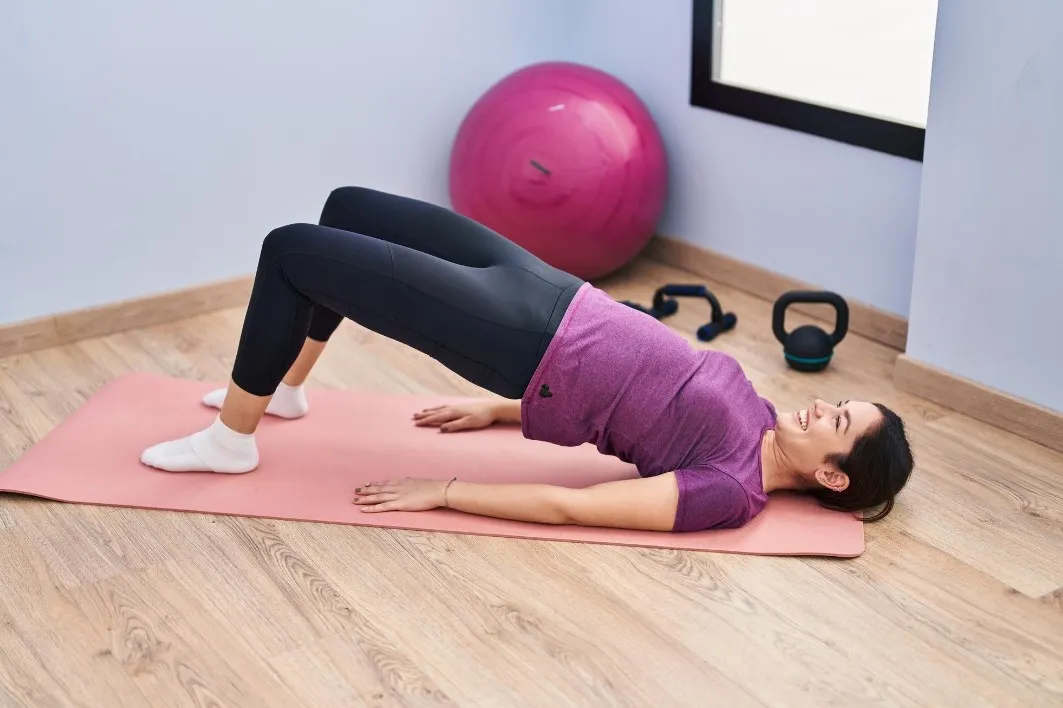 Mujer realizando ejercicio de puente sobre una colchoneta rosa en un gimnasio en casa, con una pelota de ejercicio y pesas en el fondo.