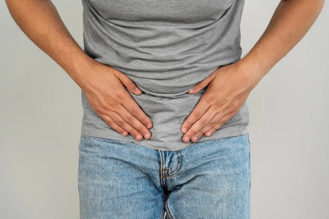 Hombre con camiseta gris sosteniendo su abdomen, mostrando signos de malestar estomacal o dolor abdominal.