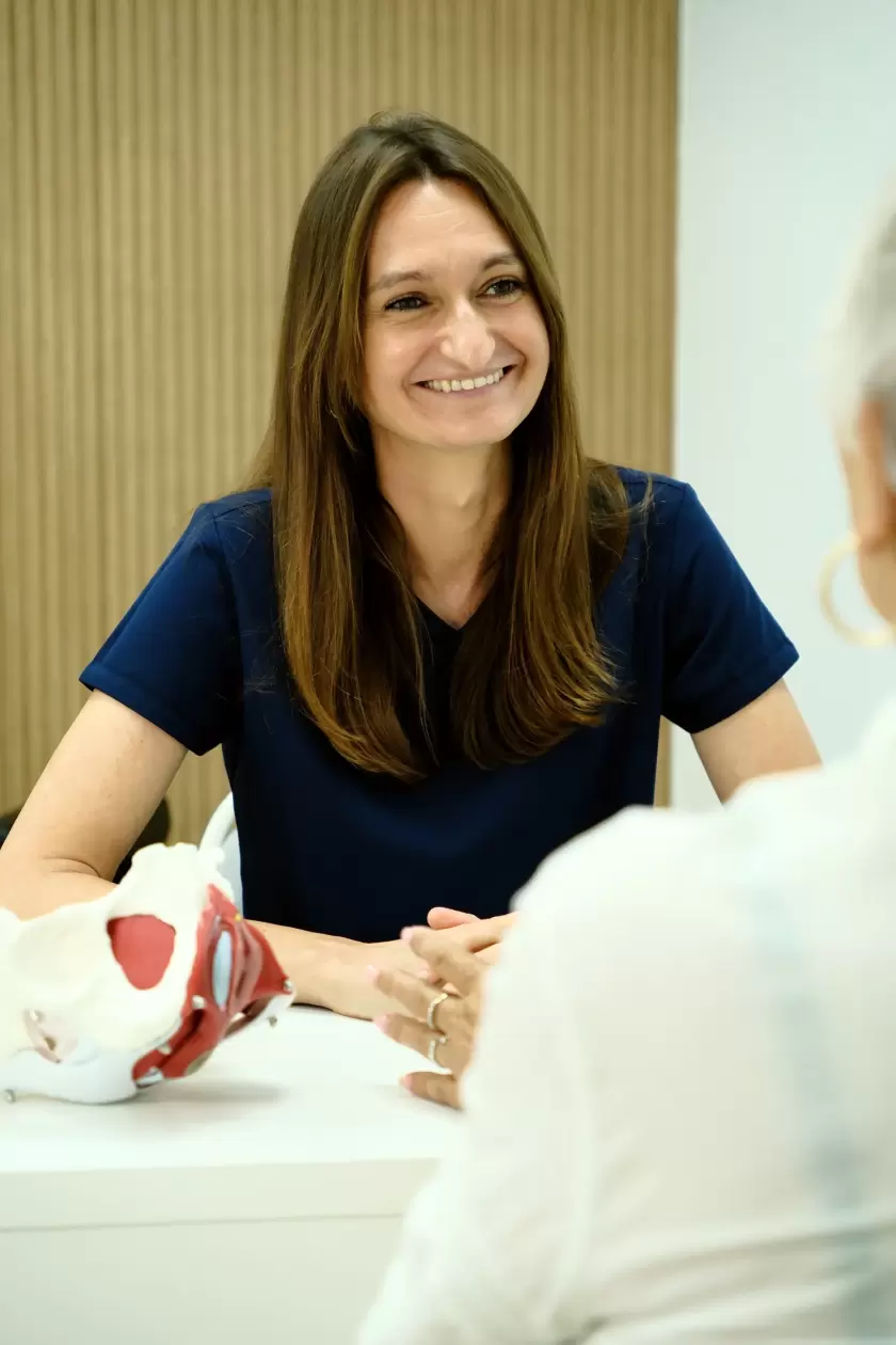 Médica sonriente en consulta, sentada frente a una paciente, con un modelo anatómico de pelvis en la mesa.
